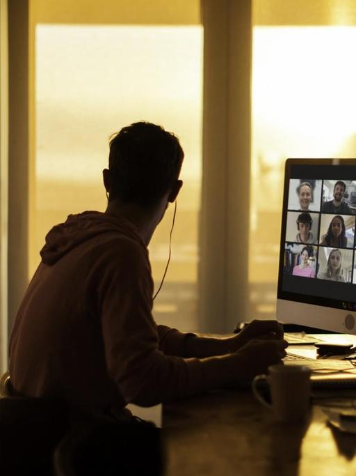 Ein junger Mann sitzt zu Hause vor einem Bildschirm mit Teilnehmern eines digitalen Treffens.
