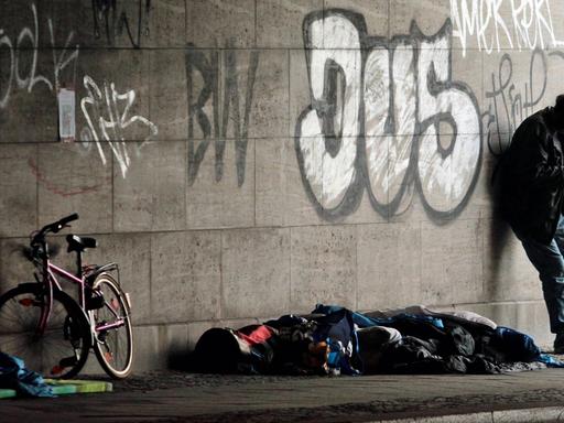 Ein obdachloser Mann steht am 9.8.2016 neben einem Schlafsack und einem Fahrrad unter einer Brücke am Bahnhof Zoo in Berlin.