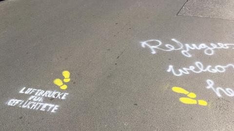 Auf den Asphalt sind gelbe Fußstapfen gemalt und mit weißer Schrift: "Luftbrücke für Geflüchtete" und "Refugees welcome here".