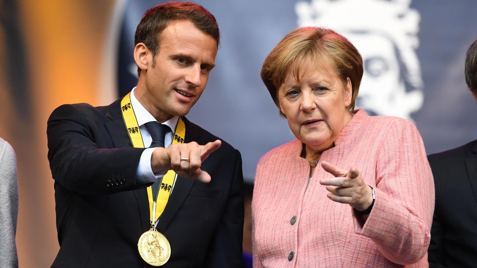 10.05.2018, Nordrhein-Westfalen, Aachen: Bundeskanzlerin Angela Merkel (CDU), und der französische Staatspräsident Emmanuel Macron stehen nach der Verleihung des Karlspreises auf einer Tribüne hinter dem Rathaus und schauen zu den Fotografen. Macron ist für seine Verdienste um die europäische Einigung in Aachen mit dem Internationalen Karlspreis ausgezeichnet worden. Foto: Ina Fassbender/dpa | Verwendung weltweit 10.05.2018, Nordrhein-Westfalen, Aachen: Bundeskanzlerin Angela Merkel (CDU), und der französische Staatspräsident Emmanuel Macron stehen nach der Verleihung des Karlspreises auf einer Tribüne hinter dem Rathaus und schauen zu den Fotografen. Macron ist für seine Verdienste um die europäische Einigung in Aachen mit dem Internationalen Karlspreis ausgezeichnet worden. Foto: Ina Fassbender/dpa | Verwendung weltweit