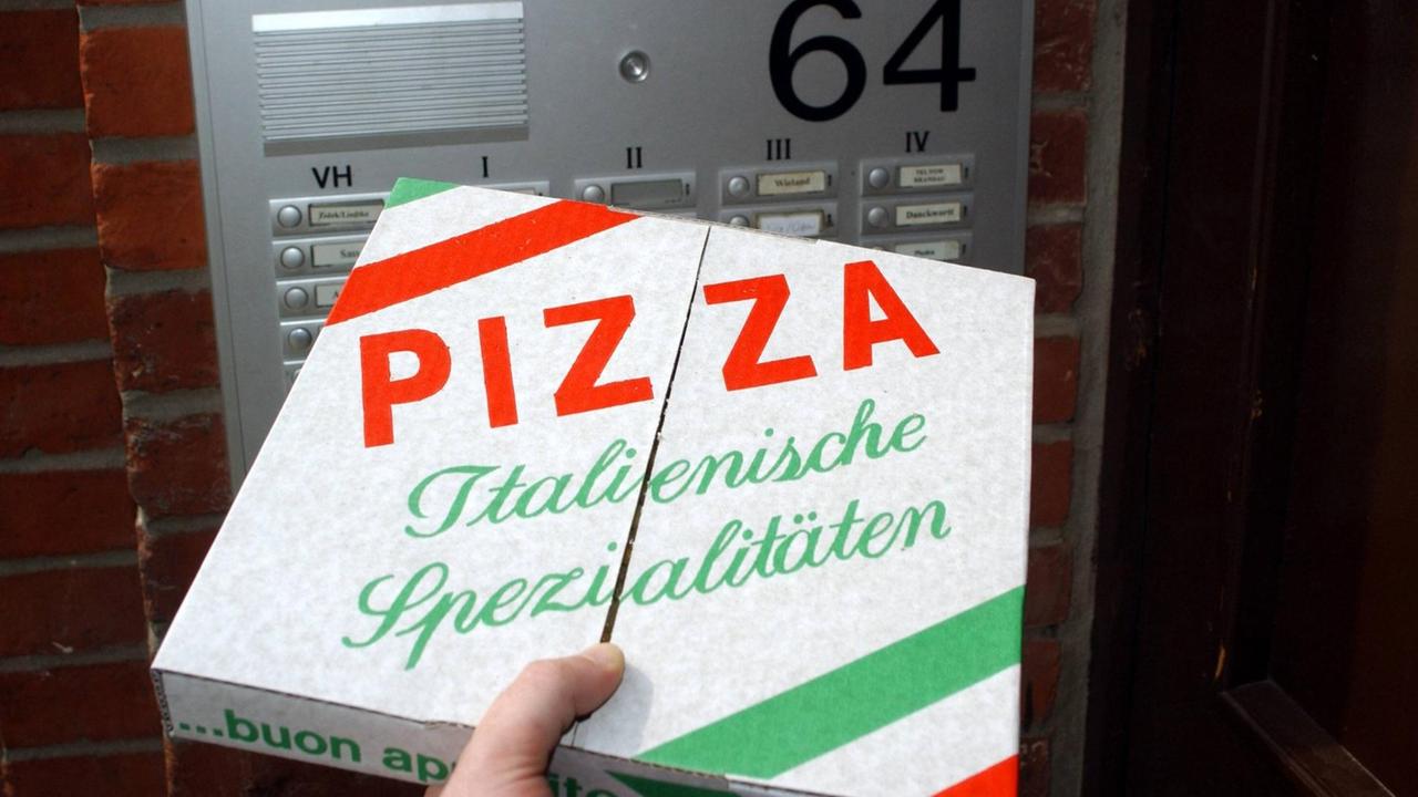 Ein Mann hält eine Pizzaschachtel in der Hand. Im Hintergrund ist die Klingelanlage eines Mehrfamilienhauses zu sehen. Ein Mann hält eine Pizzaschachtel in der Hand. Im Hintergrund ist die Klingelanlage eines Mehrfamilienhauses zu sehen.