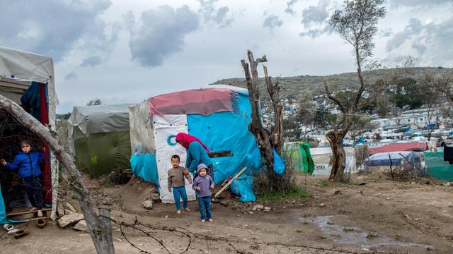 Eine Mutter mit ihren beiden Kindern in einem Flüchtlingslager auf der griechischen Insel Lesbos. Sie stehen vor einem Zelt, die Kinder sind barfuß.