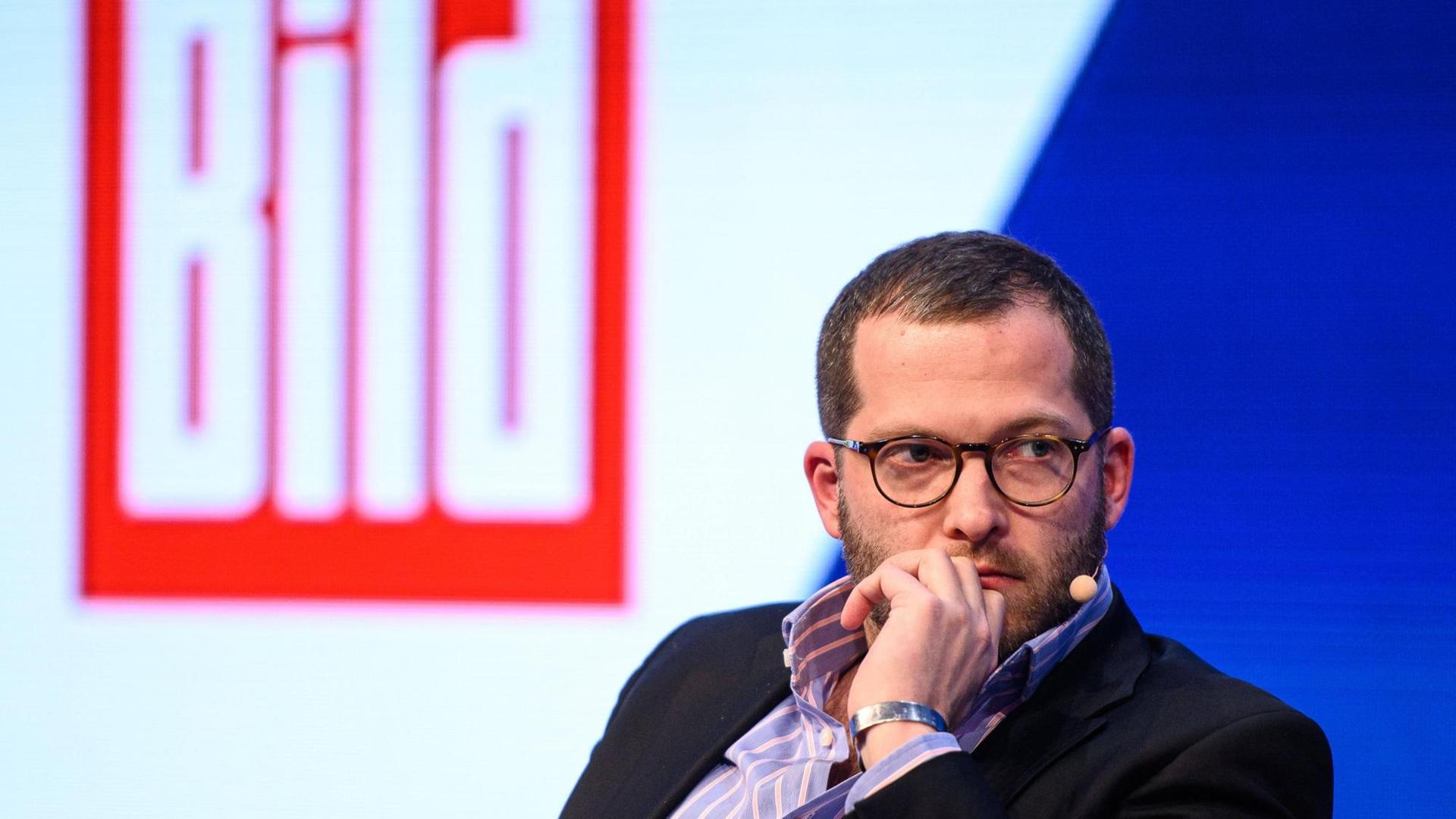 Julian Reichelt sitzt mit Brille vor dem roten Bild-Logo. Julian Reichelt sitzt mit Brille vor dem roten Bild-Logo.