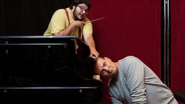 Das Bild zeigt Axel Ranisch, wie er hinter einem Klavier mit einem Dirigentenstab nach rechts zeigt, während Devid Striesow mit dem Kopf auf den Tasten des Klaviers liegt.