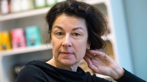 Die Schriftstellerin Zsuzsa Bánk sitzt vor einer Regalwand im Café Mathildes Kitchen in Frankfurt am Main. Sie trägt einen schwarzen Pullover und greift mit ihrer linken Hand in ihr schwarzes Haar.
