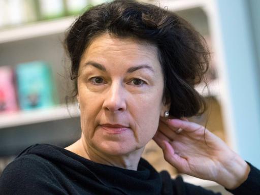 Die Schriftstellerin Zsuzsa Bánk sitzt vor einer Regalwand im Café Mathildes Kitchen in Frankfurt am Main. Sie trägt einen schwarzen Pullover und greift mit ihrer linken Hand in ihr schwarzes Haar.