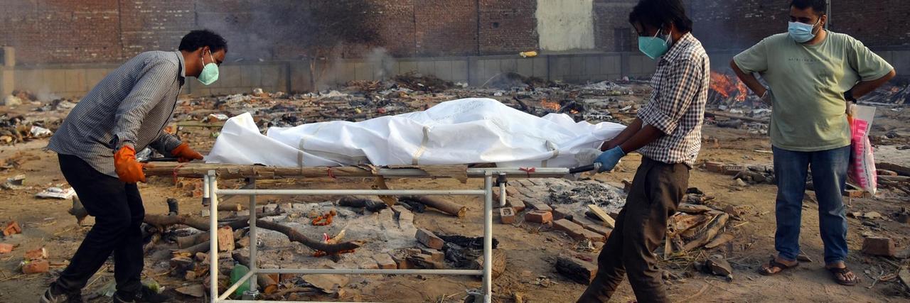 In Neu-Delhi schieben zwei Männer eine Bahre mit einem Toten an einen Platz, auf dem Leichen verbrannt werden. In Neu-Delhi schieben zwei Männer eine Bahre mit einem Toten an einen Platz, auf dem Leichen verbrannt werden.