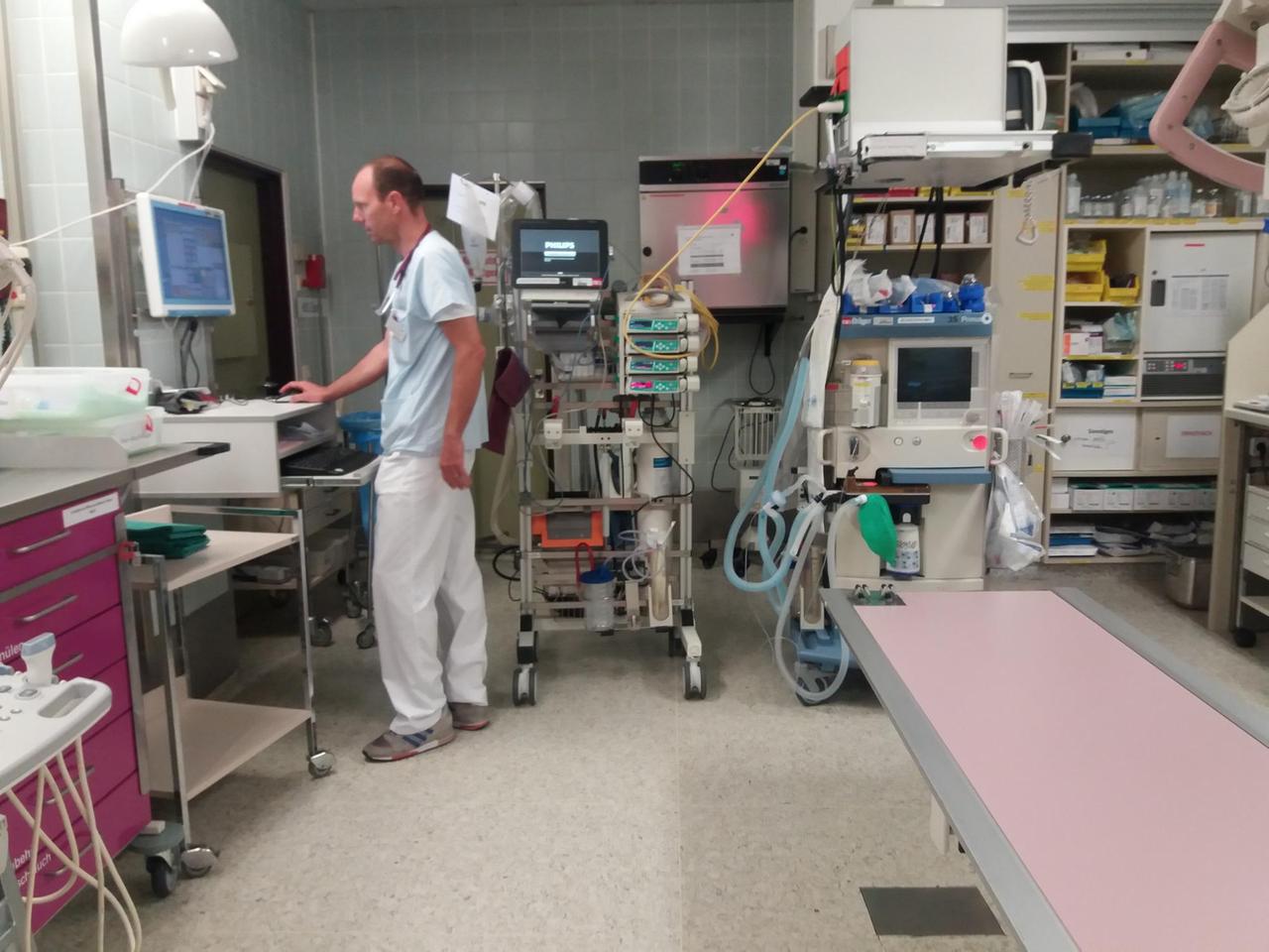 Das Bild zeigt den Oberarzt Thomas Händl in einem Schockraum der Augsburger Notaufnahme mit den verschiedensten medizinischen Gerätschaften. Hier sieht man ihn stehend an einem Computer arbeiten.