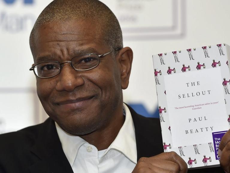 Der Afro-Amerikaner Beatty hält das Buch lächelnd neben seinen Kopf. Im Hintergrund eine Wand, auf der man unscharf die Logos des "Man Booker Prize" sieht.
