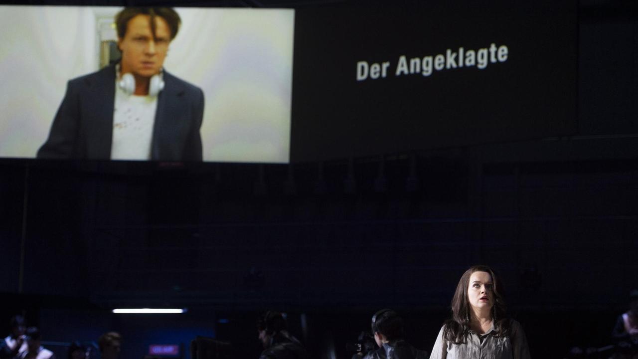 Milada steht im Vordergrund, das Bild von Dalibor ist auf einem großen Screen zu sehen mit der Aufschrift "Der Angeklagte". Milada steht im Vordergrund, das Bild von Dalibor ist auf einem großen Screen zu sehen mit der Aufschrift "Der Angeklagte".
