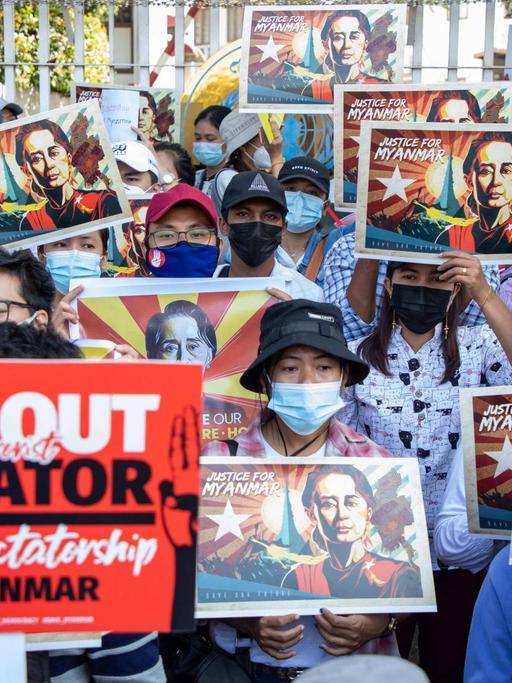 Junge Demonstrierende in Myanmar halten Schilder mit dem Porträt von Aung San Suu Kyi hoch. Ein Demonstrant hält ein rotes Schild mit der Aufschrift "Get out Dictator".