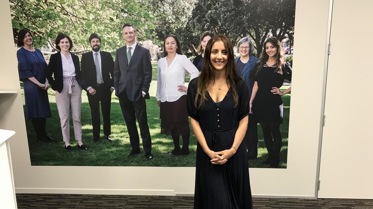 Eine junge Frau mit langen dunklen Haaren und schwarzem Kleid posiert vor einem Foto mit mehreren Personen im Grünen. Eine junge Frau mit langen dunklen Haaren und schwarzem Kleid posiert vor einem Foto mit mehreren Personen im Grünen.