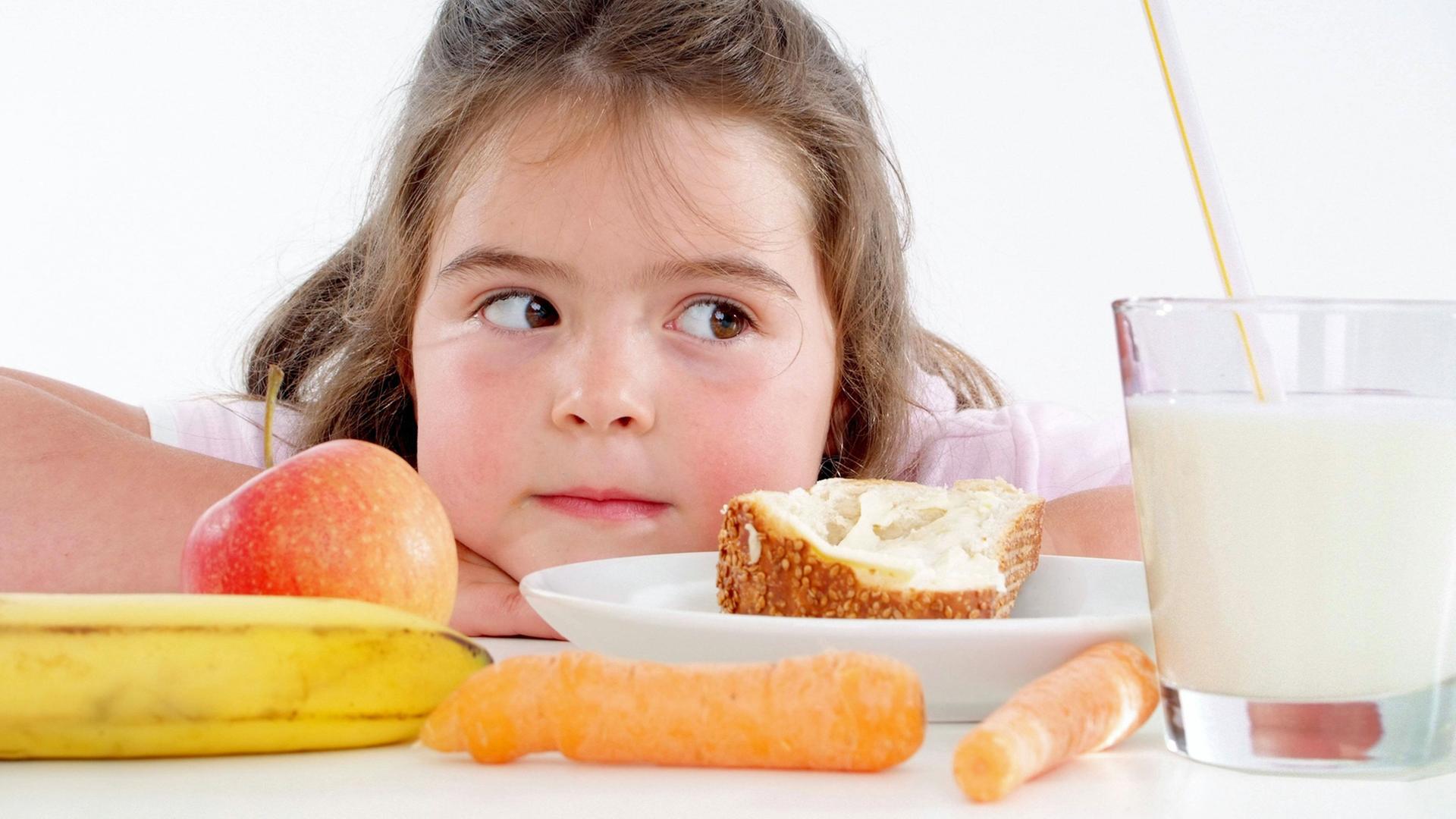 Ein Mädchen sitzt vor Obst, Gemüse, Brot und Milch - Lebensmittel die gemeinhin als gesund gelten Ein Mädchen sitzt vor Obst, Gemüse, Brot und Milch - Lebensmittel die gemeinhin als gesund gelten