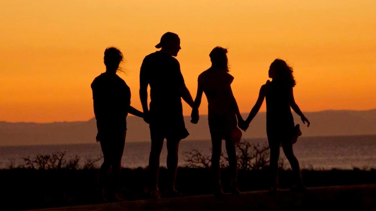 Eine aus vier Personen bestehende Familie steht an einem Strand im Dämmerlicht, sodass nur ihre Silhouetten zu sehen sind. Eine aus vier Personen bestehende Familie steht an einem Strand im Dämmerlicht, sodass nur ihre Silhouetten zu sehen sind.