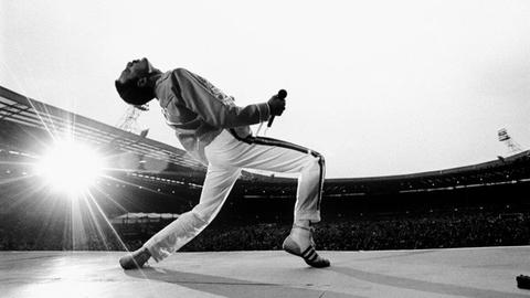 "Queen"-Sänger Freddie Mercury bei einem Konzert im Wembley Stadium. Der Sänger hat den Rücken nach hinten gebeugt, hinter ihm ist eine riesige Menschenmenge.