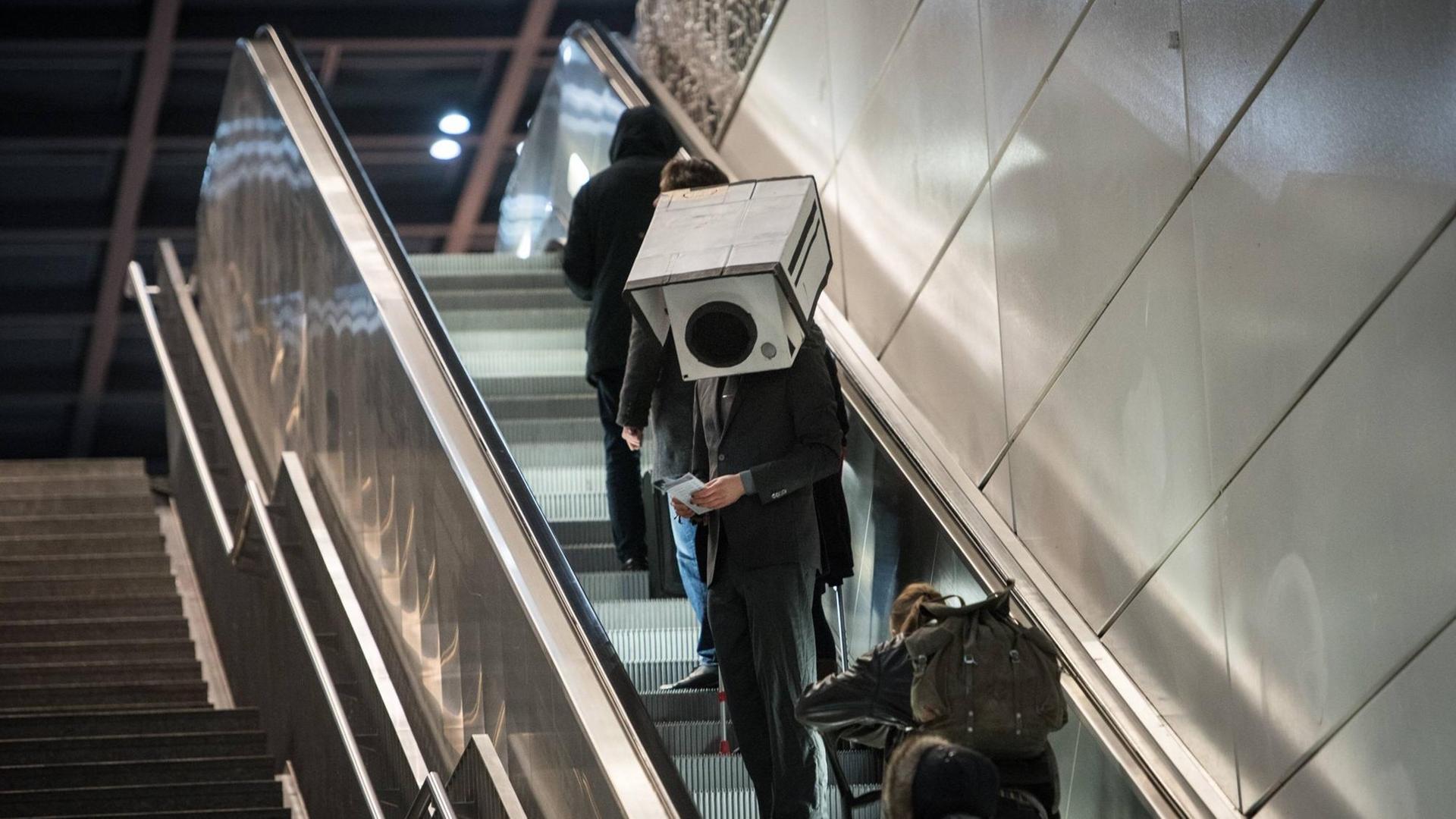 Ein Mann im Anzung und mit einer Überwachungskamera aus Pappe auf dem Kopf fährt eine Rollpreppe hoch. Ein Mann im Anzung und mit einer Überwachungskamera aus Pappe auf dem Kopf fährt eine Rollpreppe hoch.