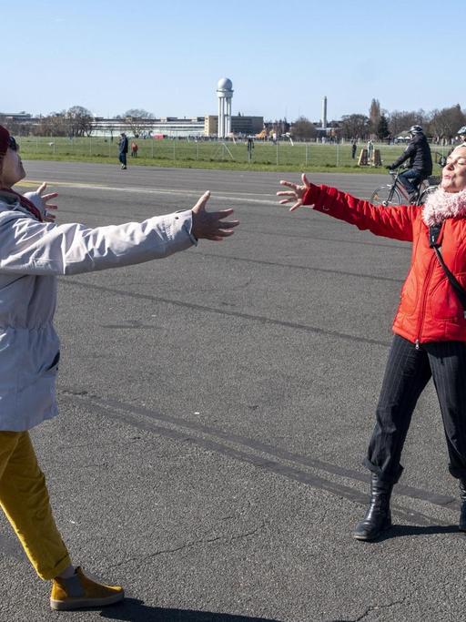 Zwei Frauen Begrüßen sich mit einer angedeuteten Umarmung aus der Distanz auf dem Tempelhofes Feld in Berlin.