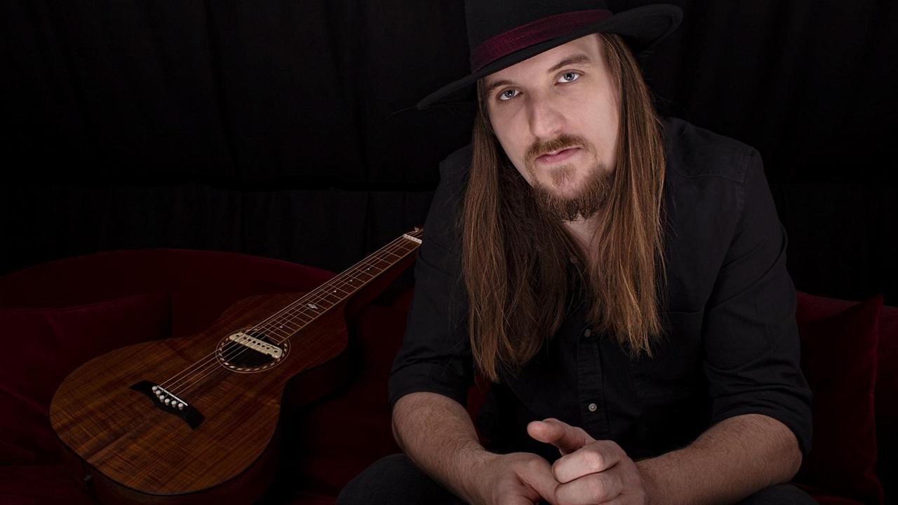Ein großer Mann mit langen, dunklen Haaren und Hut auf dem Kopf blickt in die Kamera. Neben ihm liegt eine Gitarre.