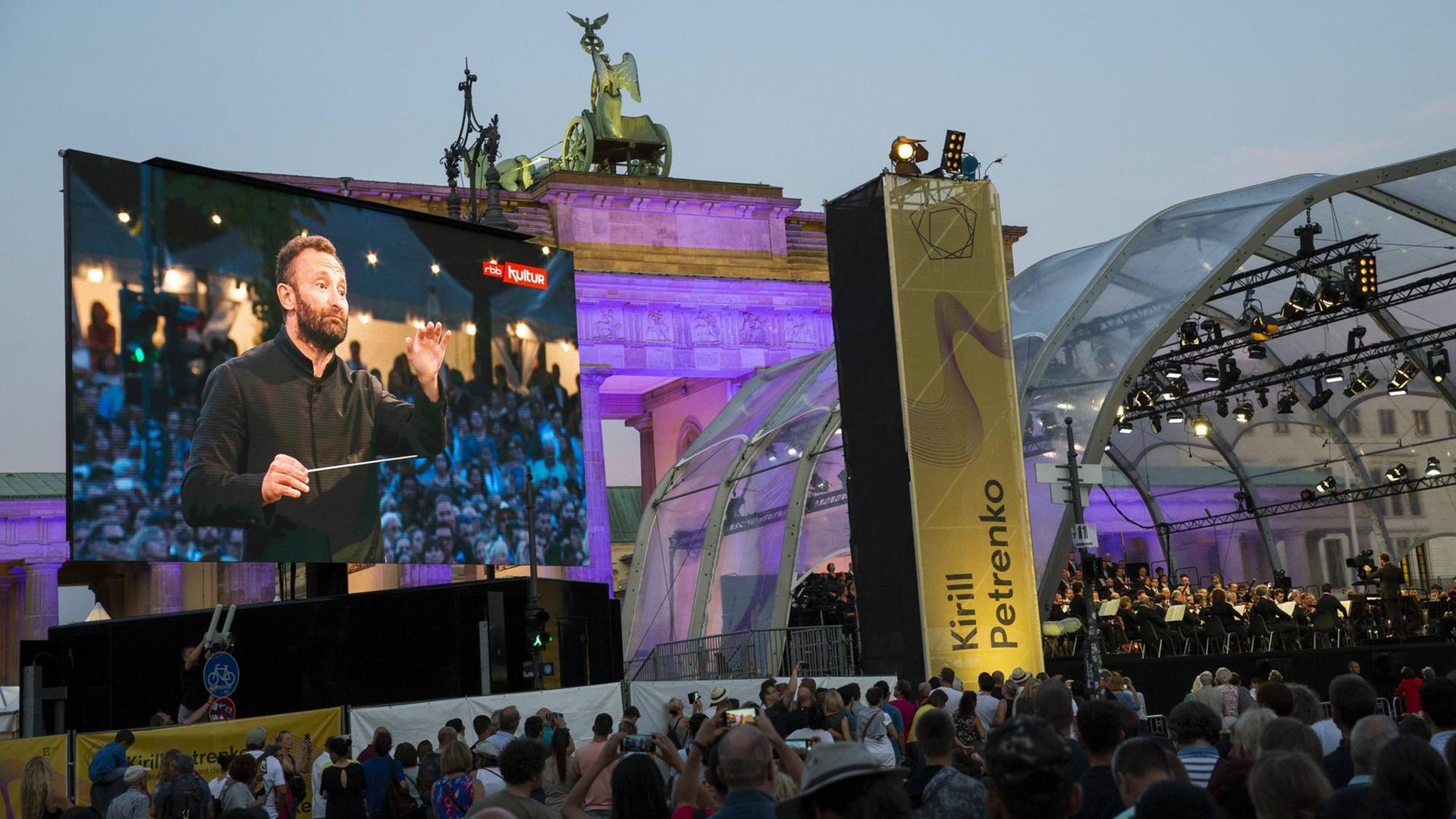 Kirill Petrenko ist auf einer großen Leinwand dabei zu sehen, wie er die Berliner Philharmoniker dirigiert, im Hintergrund das Brandenburger Tor. Kirill Petrenko ist auf einer großen Leinwand dabei zu sehen, wie er die Berliner Philharmoniker dirigiert, im Hintergrund das Brandenburger Tor.