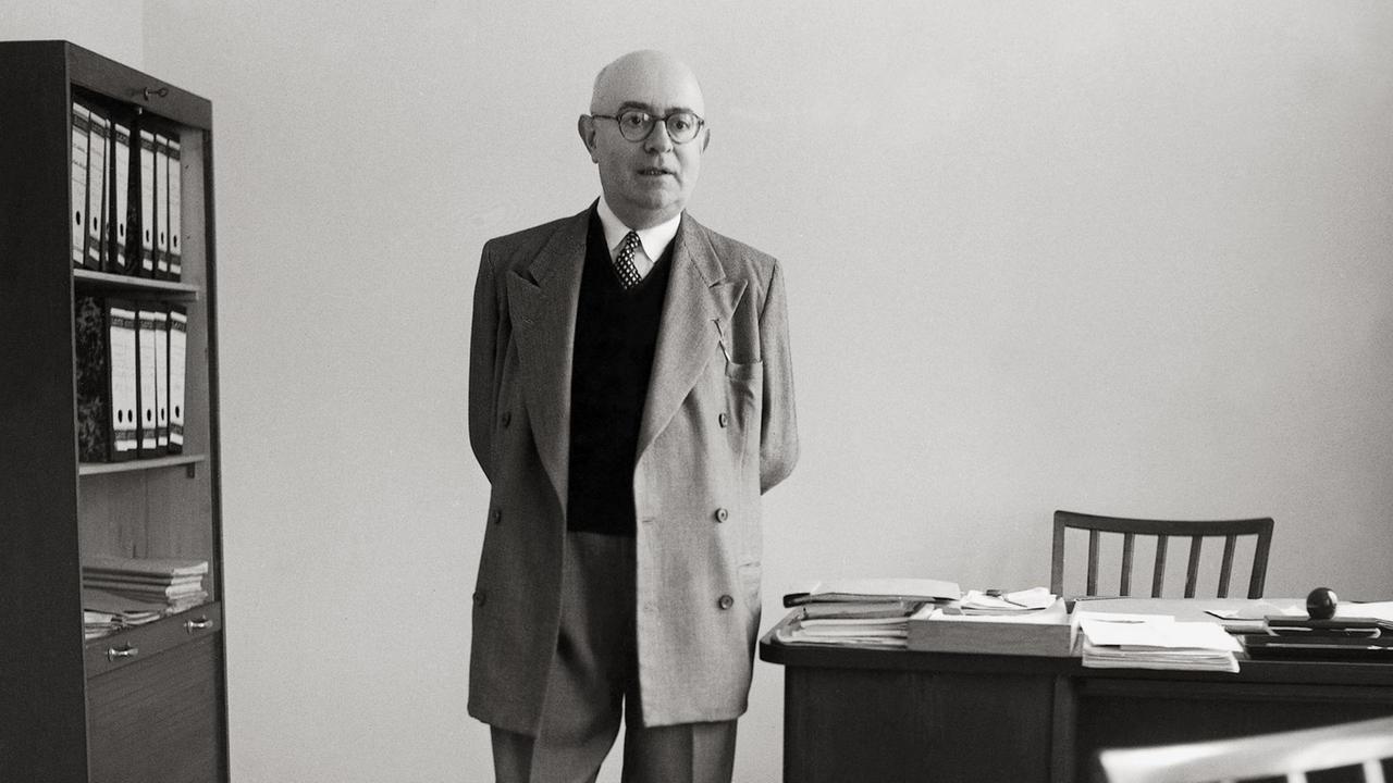 Die Aufnahme von 1958 zeigt den Philosophen Theodor W. Adorno mit hinter dem Rücken verschränkten Händen neben seinem Schreibtisch stehend.
