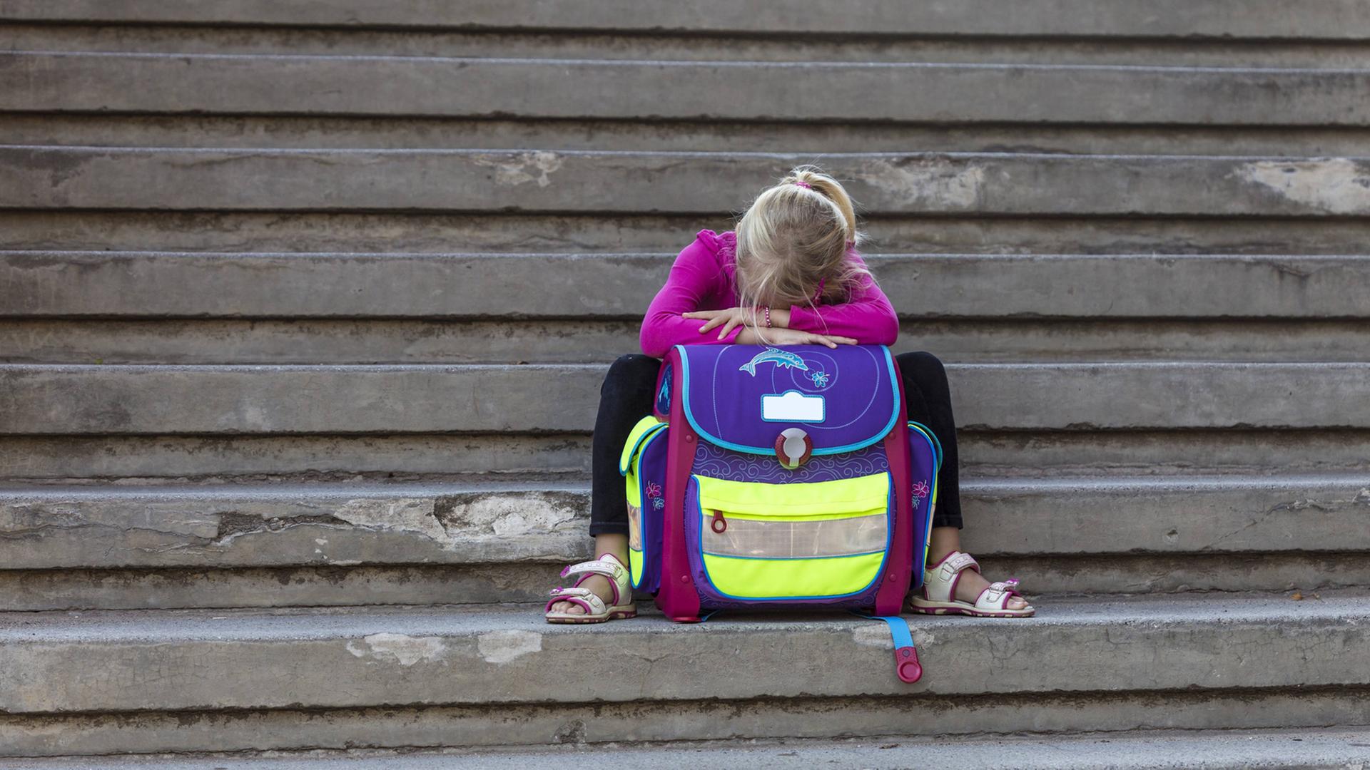 Ein Mädchen sitzt traurig auf Steinstufen, den Kopf auf den Tornister gelehnt. Ein Mädchen sitzt traurig auf Steinstufen, den Kopf auf den Tornister gelehnt.