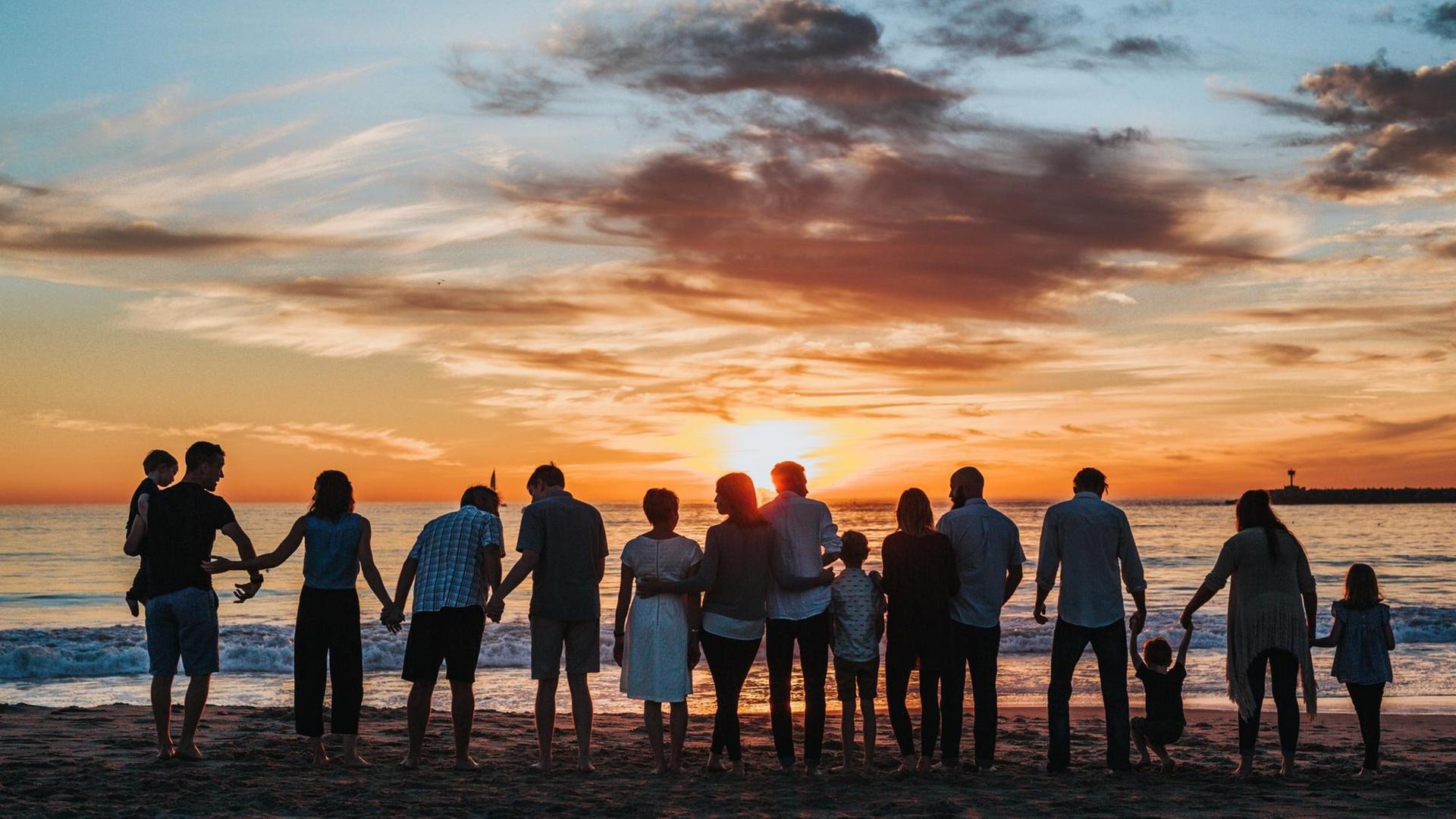Eine Großfamilie mit mehreren Kindern steht am Strand mit den Rücken zur Kamera und betrachtet den Sonnenuntergang.