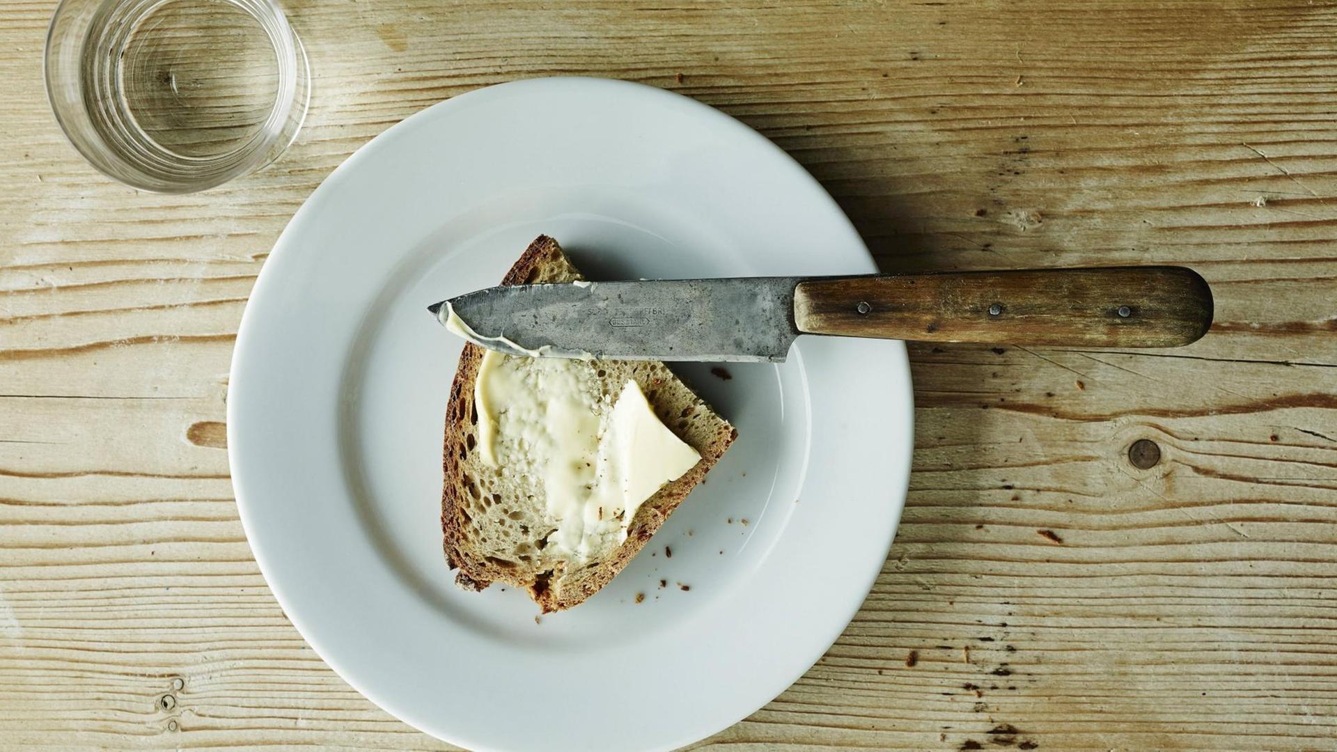 Auf einem Tisch stehen ein Wasserglas und eine Scheibe Brot mit Butter. Auf einem Tisch stehen ein Wasserglas und eine Scheibe Brot mit Butter.
