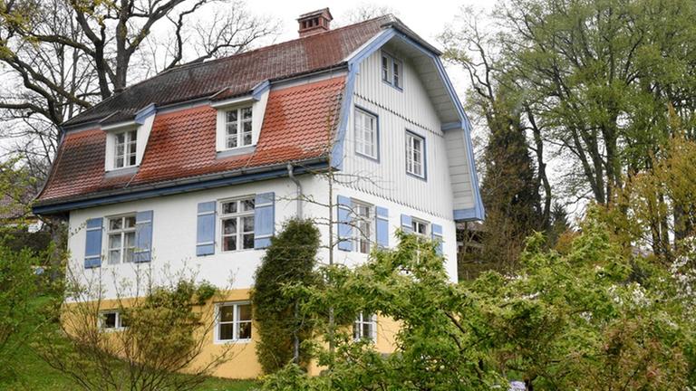 Ein großes, weißes Haus mit rotem Dach und blauen Fensterläden steht in Hanglage von Bäumen umgeben. Ein großes, weißes Haus mit rotem Dach und blauen Fensterläden steht in Hanglage von Bäumen umgeben.