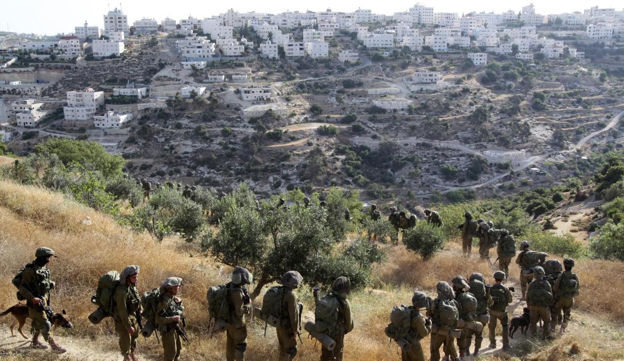 Vor dem Hintergrund Hebrons laufen zahlreiche israelische Soldaten im Gänsemarsch einen Pfad an einem Hang hinunter.