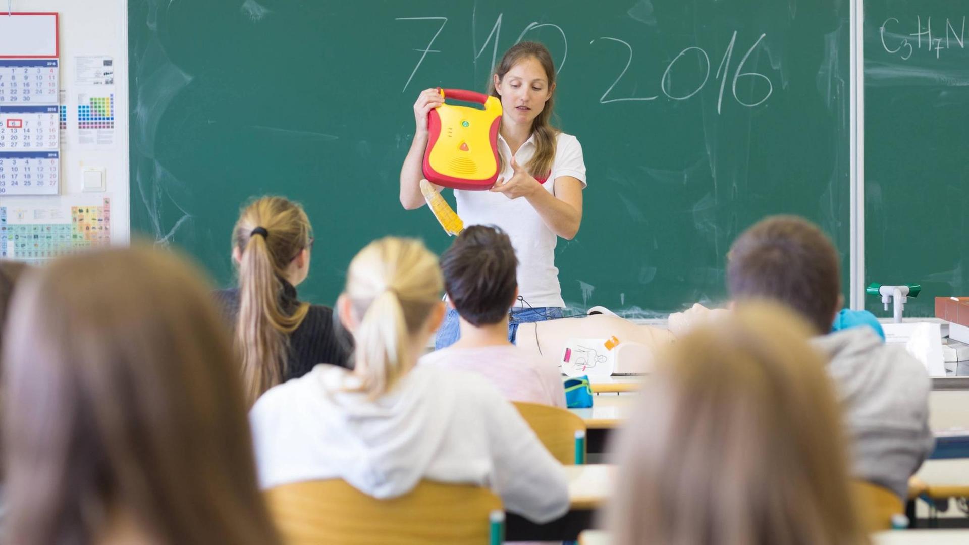 Ein Frau hält vor einer Schulklasse einen gelb-roten Kasten in die Höhe.