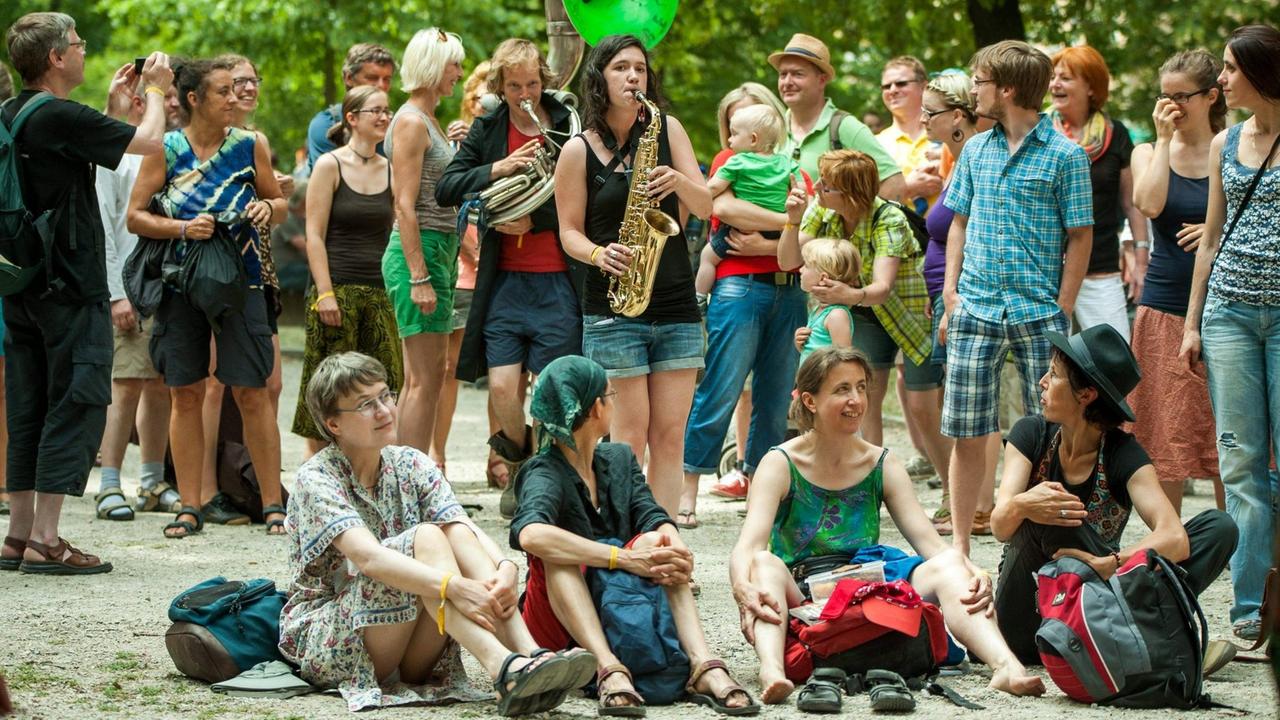Zwei Musikerinnen sind von buntem Publikum in sommerlicher Kleidung stehend und sitzend umringt. Zwei Musikerinnen sind von buntem Publikum in sommerlicher Kleidung stehend und sitzend umringt.