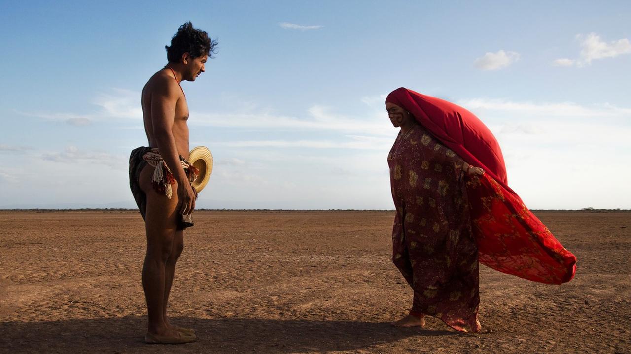 Ein Mann mit nakten Oberkörper und eine Frau mit roten, langen Kopftuch stehen sich in der Wüste gegenüber.