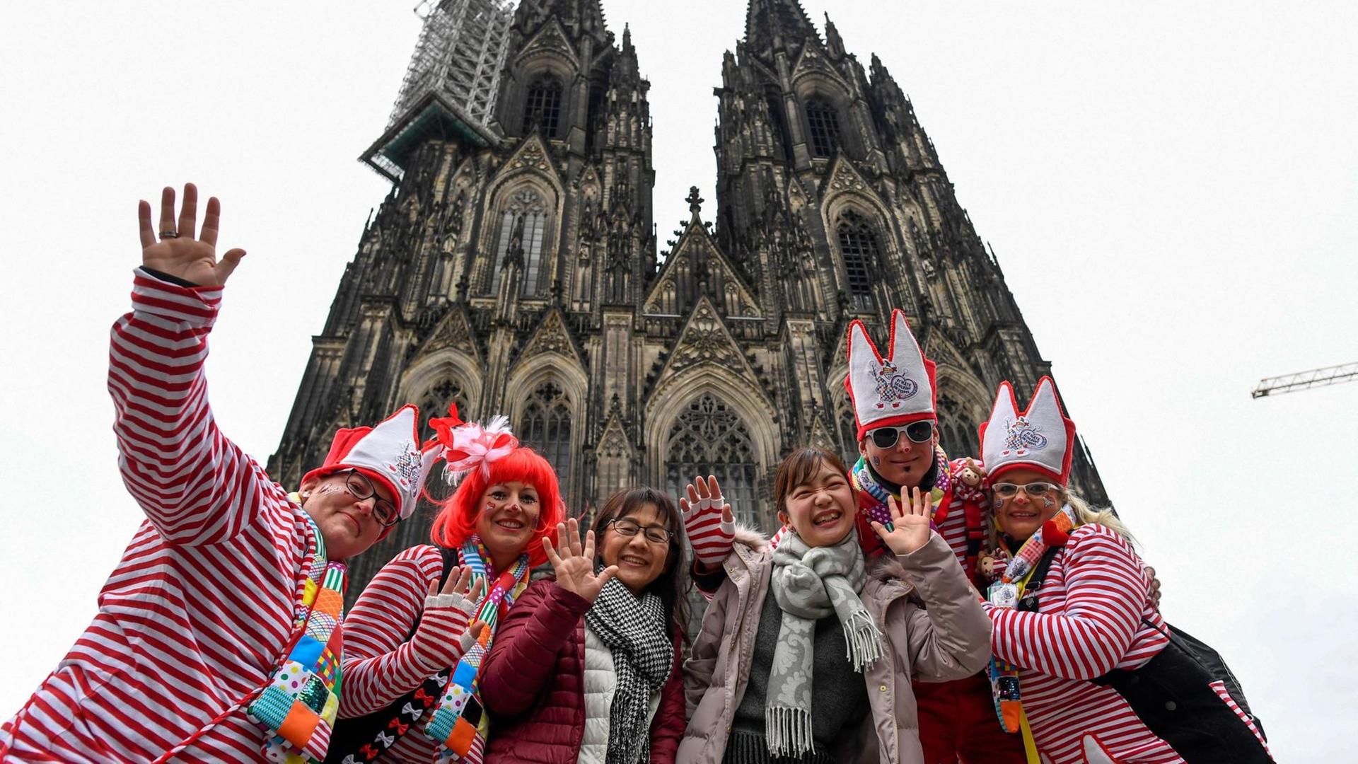 Eine Gruppe verkleideter Karnevalisten nimmt vor dem Kölner Dom zwei japanische Touristinnen in den Arm