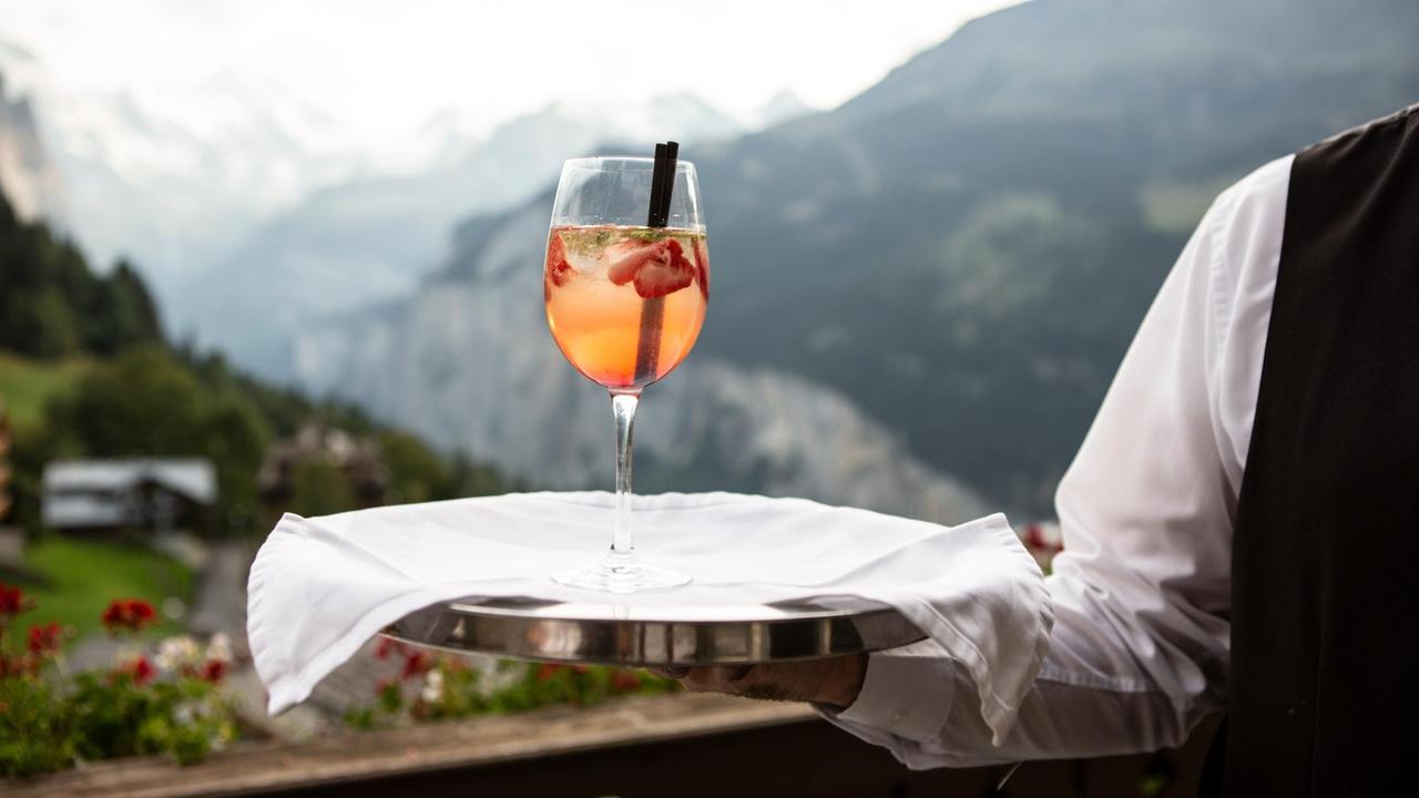 Ein Kellner serviert einen Drink auf dem Silbertablett vor idyllischer Bergkulisse. Ein Kellner serviert einen Drink auf dem Silbertablett vor idyllischer Bergkulisse.