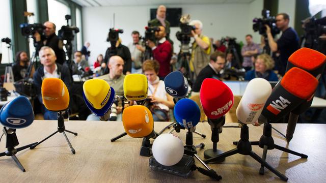 Auf einem Tisch stehen ein Dutzend Mikrofone, im Hintergrund sind Journalisten, zum Teil mit Kameras zu sehen.