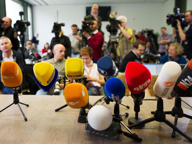 Auf einem Tisch stehen ein Dutzend Mikrofone, im Hintergrund sind Journalisten, zum Teil mit Kameras zu sehen.