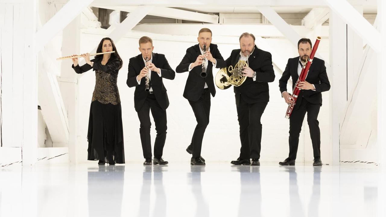 Das Ensemble steht in einem weißen Raum und scheint sich an die Kamera heranzuschleichen, während sie ihre Instrumente spielen. Das Ensemble steht in einem weißen Raum und scheint sich an die Kamera heranzuschleichen, während sie ihre Instrumente spielen.