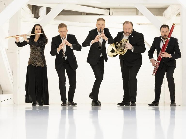 Das Ensemble steht in einem weißen Raum und scheint sich an die Kamera heranzuschleichen, während sie ihre Instrumente spielen.