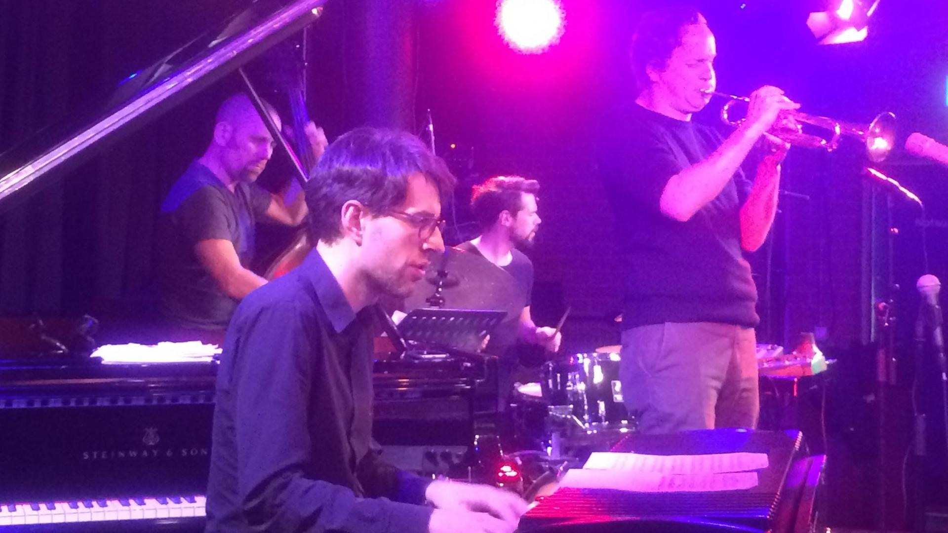 Ein Konzertbild in violett-roter Bühnenbeleuchtung. Hinter einem Pianisten im Vordergrund ist rechts ein Trompeter zu sehen, links spielt ein Kontrabassist und hinten mittig ein Schlagzeuger. Ein Konzertbild in violett-roter Bühnenbeleuchtung. Hinter einem Pianisten im Vordergrund ist rechts ein Trompeter zu sehen, links spielt ein Kontrabassist und hinten mittig ein Schlagzeuger.