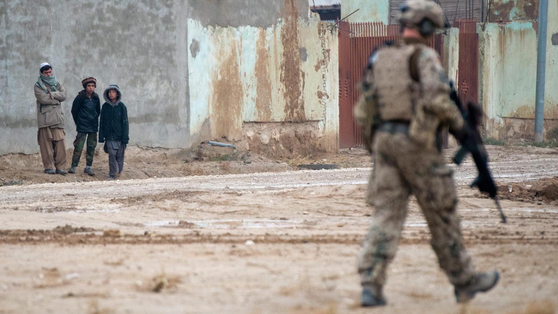 Straßenszene aus Masar-i-Scharif in Afghanistan: Ein deutscher Soldat patroulliert durch den Vordergrund des Bildes, mit dem Rücken zum Fotografen. Er bewegt sich weg von drei afghanischen Zivilisten, die im Hintergrund des Bildes stehen und zu ihm schauen. Straßenszene aus Masar-i-Scharif in Afghanistan: Ein deutscher Soldat patroulliert durch den Vordergrund des Bildes, mit dem Rücken zum Fotografen. Er bewegt sich weg von drei afghanischen Zivilisten, die im Hintergrund des Bildes stehen und zu ihm schauen.