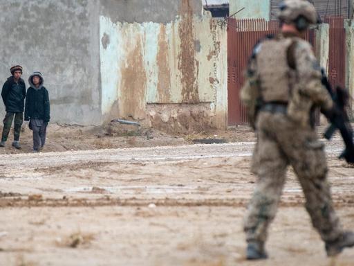 Straßenszene aus Masar-i-Scharif in Afghanistan: Ein deutscher Soldat patroulliert durch den Vordergrund des Bildes, mit dem Rücken zum Fotografen. Er bewegt sich weg von drei afghanischen Zivilisten, die im Hintergrund des Bildes stehen und zu ihm schauen.