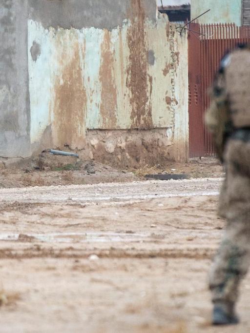 Straßenszene aus Masar-i-Scharif in Afghanistan: Ein deutscher Soldat patroulliert durch den Vordergrund des Bildes, mit dem Rücken zum Fotografen. Er bewegt sich weg von drei afghanischen Zivilisten, die im Hintergrund des Bildes stehen und zu ihm schauen.