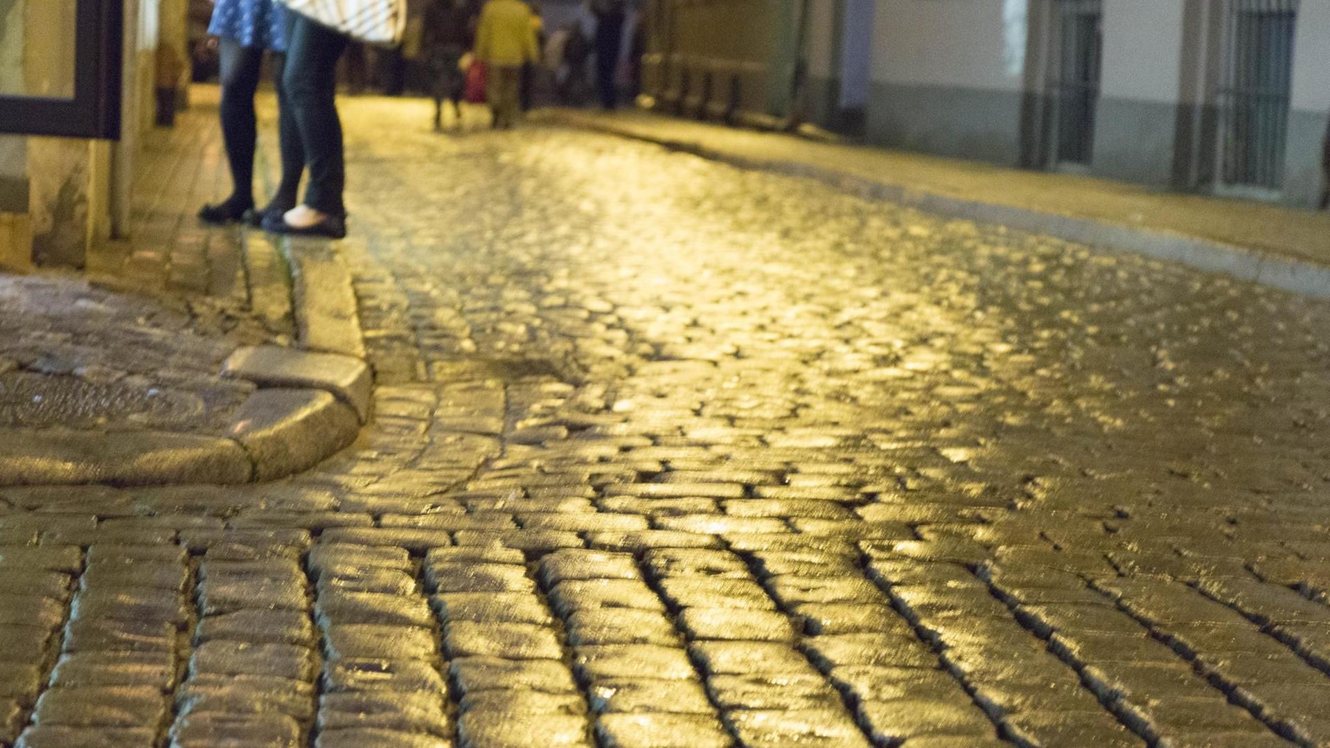 Passanten auf nassem Kopfsteinpflaster an einer Straßenecke in der Nacht.