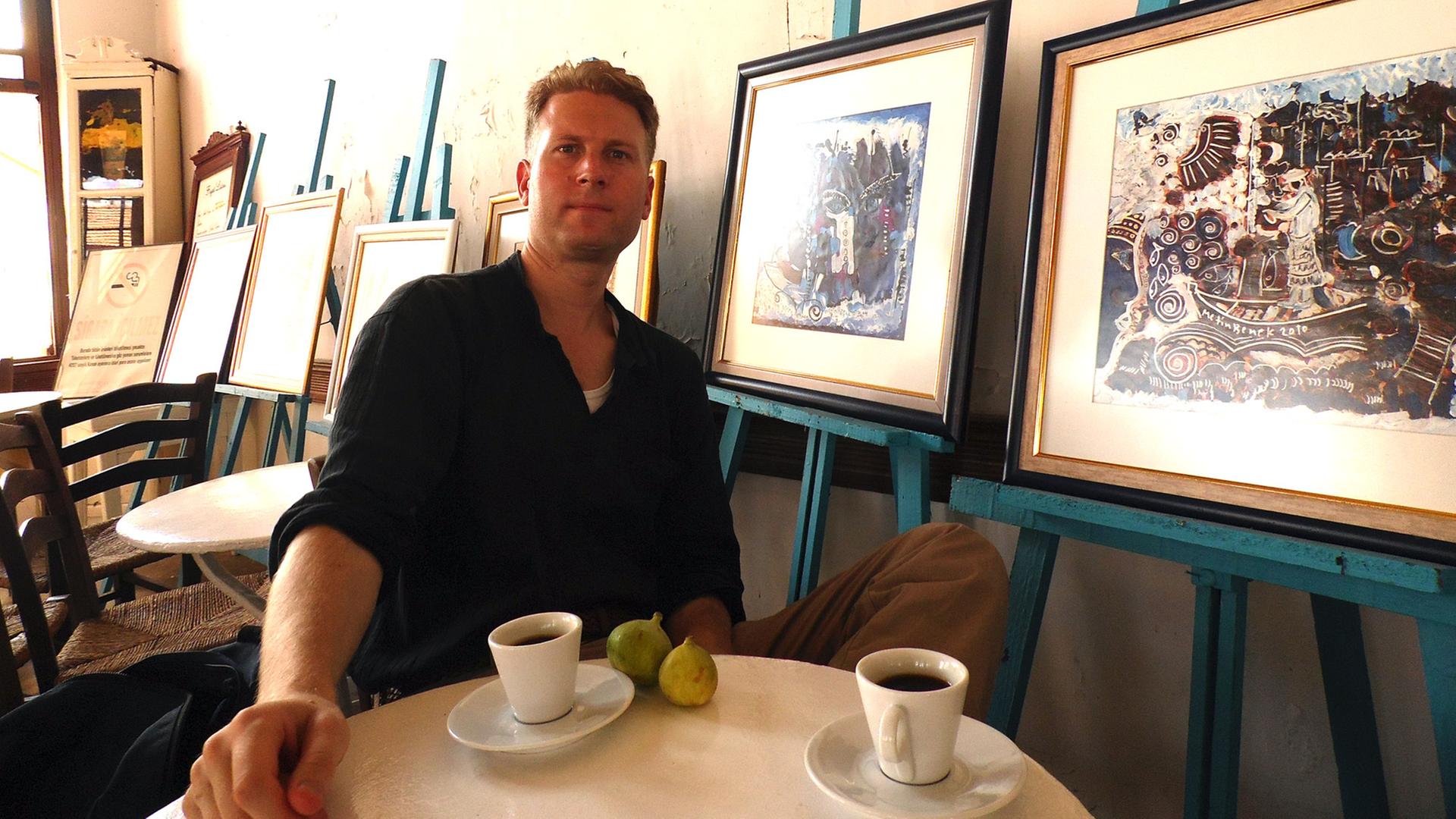 Ein Mann sitzt an einem Tisch vor Kaffeetassen, an der Wand lehnen Bilder.