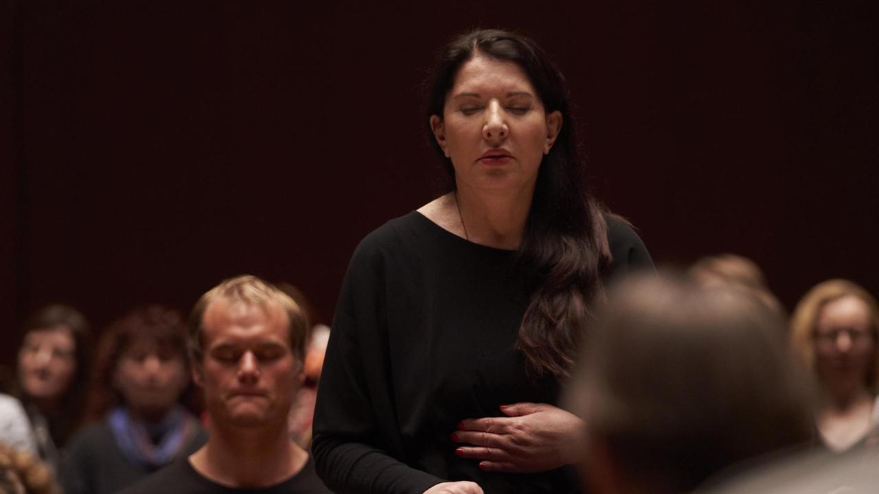 Marina Abramovic steht im Publikum, Hand auf dem Bauch und Augen geschlossen. Sie trägt schwarz. Marina Abramovic steht im Publikum, Hand auf dem Bauch und Augen geschlossen. Sie trägt schwarz.