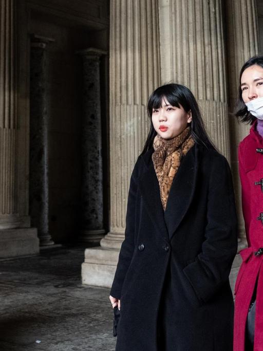 Zwei junge Chinesinnen laufen in Wintermänteln an historischen Gebäuden vorbei, eine von ihnen trägt eine Atemschutzmaske.