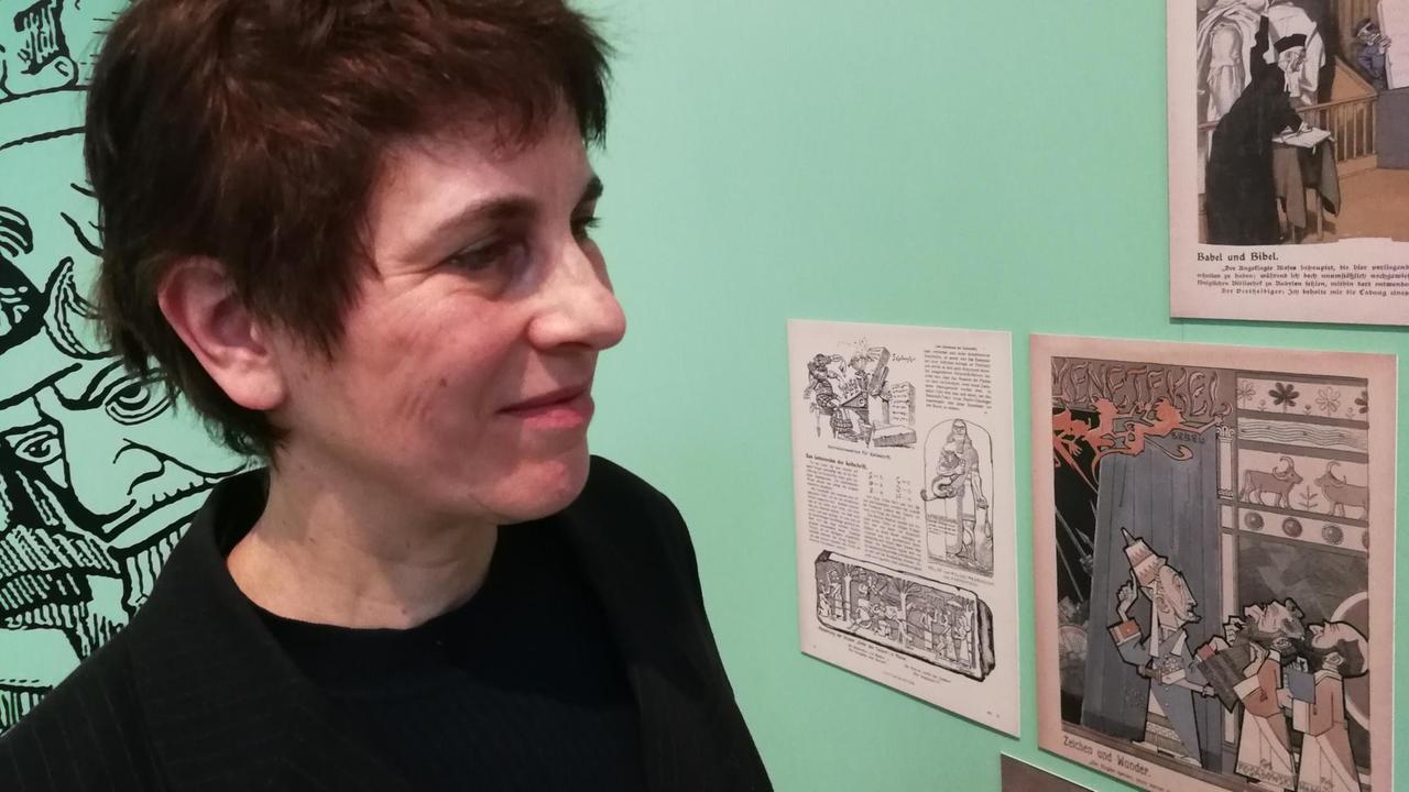 Die Altorientalistin Eva Cancik-Kirschbaum steht vor einer Wand, mit mehreren Aushängen, darunter eine Karikatur mit Figuren im assyrischen Stil Die Altorientalistin Eva Cancik-Kirschbaum steht vor einer Wand, mit mehreren Aushängen, darunter eine Karikatur mit Figuren im assyrischen Stil