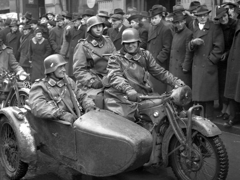 Zwei Soldaten auf dem Motorrad, einer im Beiwagen, fahren durch eine von Menschen gesäumte Straße in Prag
