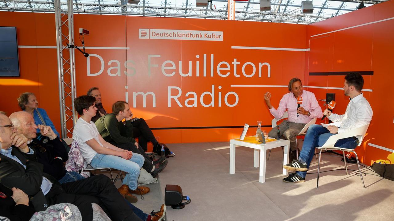 Links im Bild ist das Publikum zu sehen, dass auf Stühlen vor dem Gesprächsgast und dem Moderator sitzt. Links im Bild ist das Publikum zu sehen, dass auf Stühlen vor dem Gesprächsgast und dem Moderator sitzt.