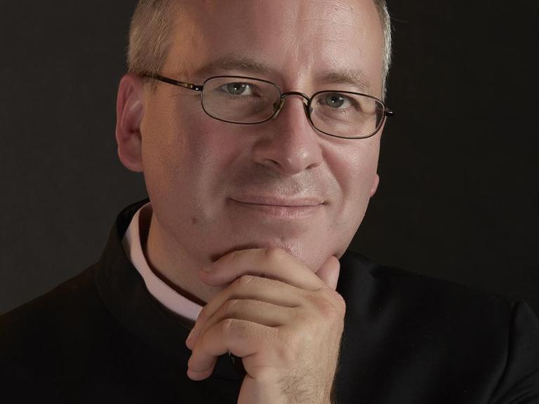 Ein Mann mit Brille schaut in die Kamera, wobei er eine Hand an sein Kinn gelegt hat.